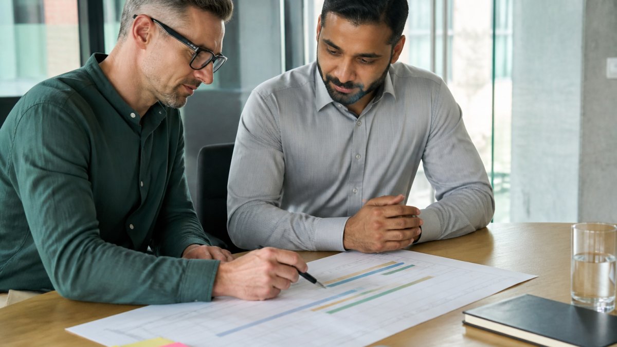 A project manager and client reviewing a printed project timeline on a table in a bright modern office, laptop and sticky notes nearby
