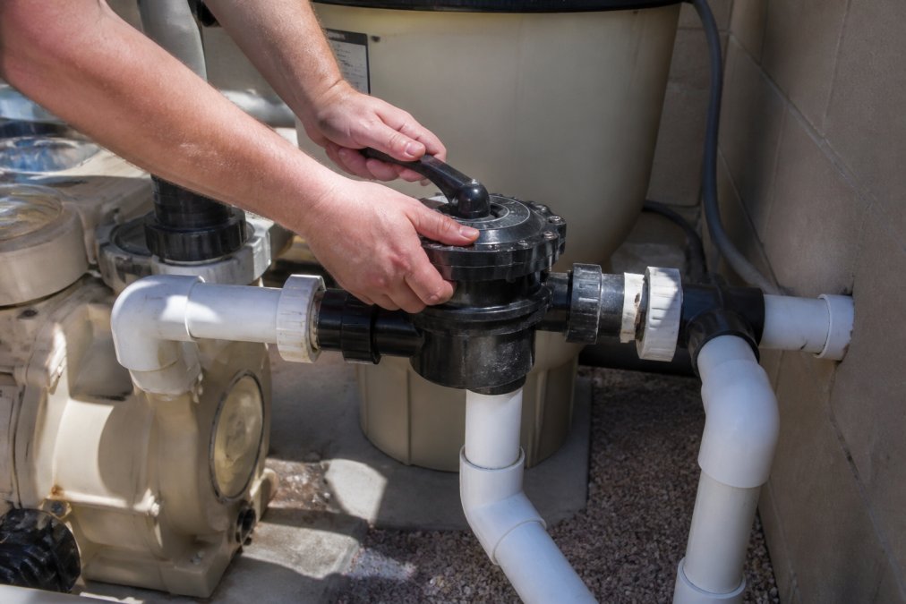 Close-up of a pool equipment pad showing a diverter valve and plumbing connected to a solar heating system, with a technician's hands adjusting the valve in bright daylight
