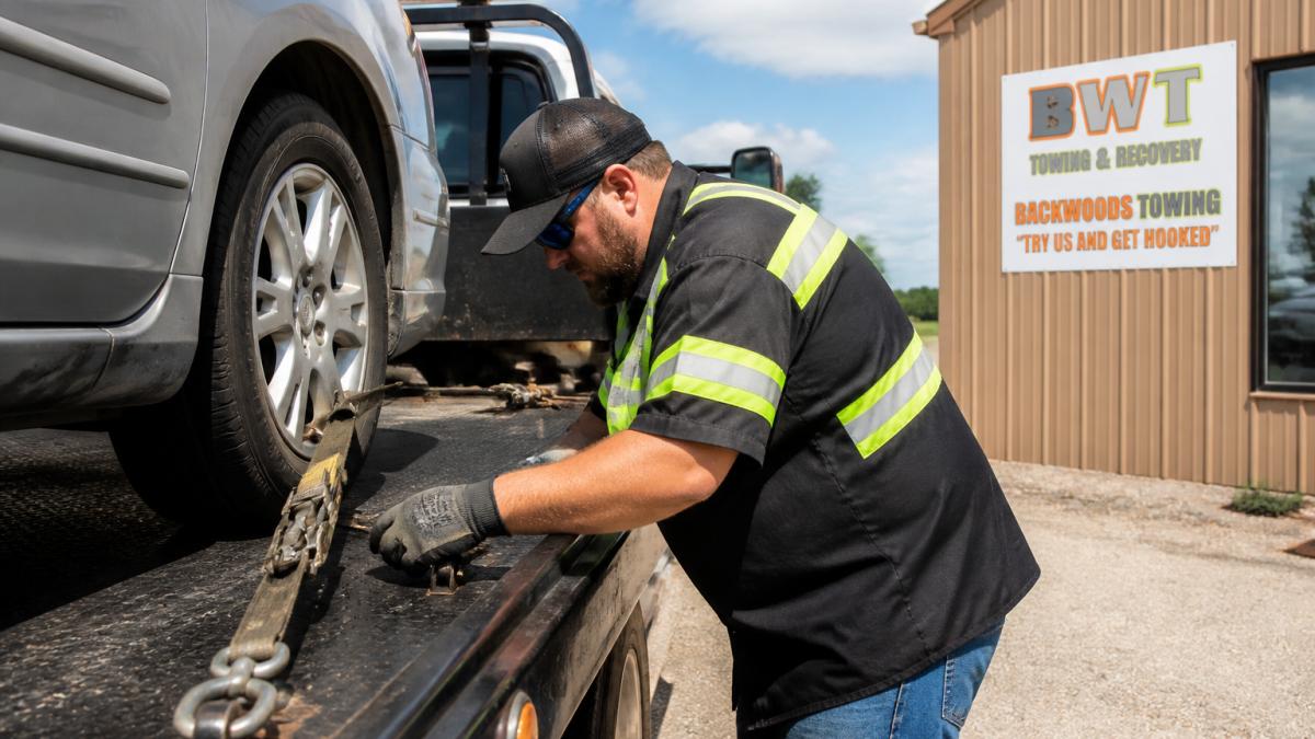 Professional tow truck operator securing a vehicle on the flatbed with heavy-duty straps and chains, showing proper equipment handling and vehicle positioning during daytime operation