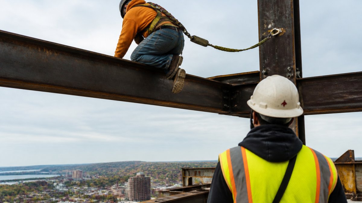A construction worker wearing a full-body harness and lanyard, secured to an anchor point while working on an elevated steel beam, with a safety monitor observing nearby