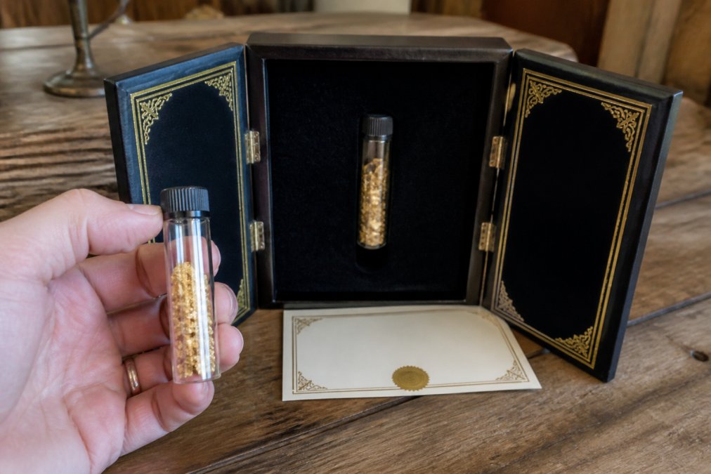 A hand holding a small glass vial filled with raw gold flakes beside an open handcrafted tri-fold display case with embossed gold detailing and a Certificate of Authenticity on a wooden table