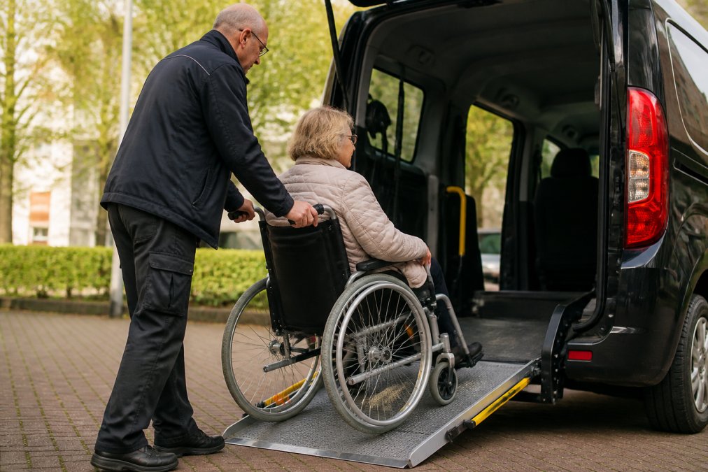 Rolstoelgebruiker die voorzichtig in een aangepaste rolstoeltaxi stapt met hulp van een ervaren chauffeur, waarbij de oprijplaat zichtbaar is en natuurlijk daglicht de scène verlicht