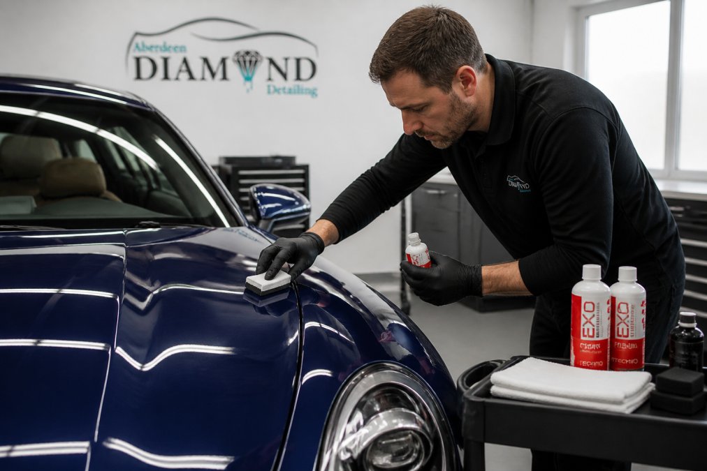 A professional detailer wearing nitrile gloves applying a ceramic coating to the deep blue paintwork of a luxury car inside a brightly lit detailing studio, with coating bottles and applicator pads visible on a nearby trolley