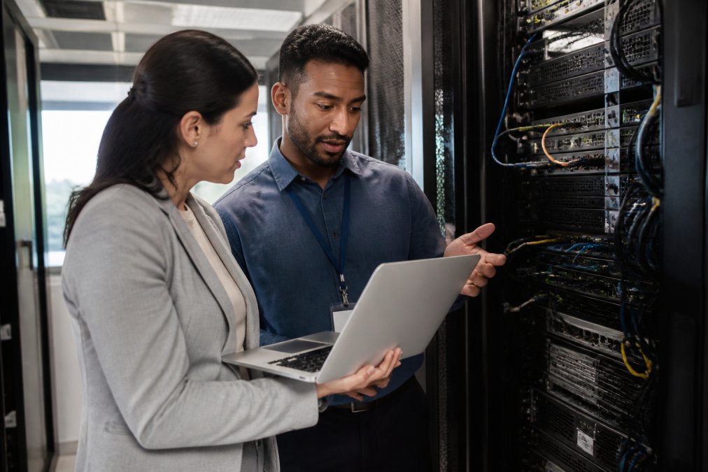 A business owner and an IT manager reviewing a server rack in a modern office, discussing data protection plans with a laptop open showing a dashboard