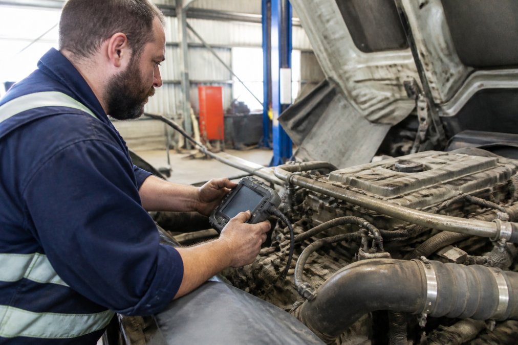 Professional mechanic using modern diagnostic scanner on diesel truck engine in bright workshop with specialized equipment visible
