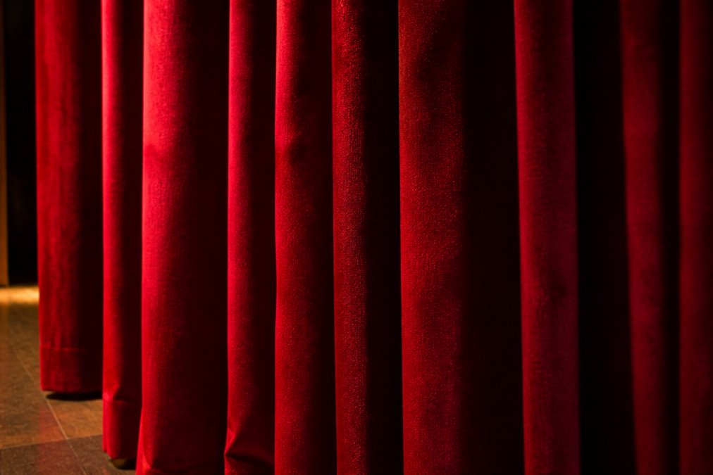 Close-up photograph of rich, plush IFR velour stage curtains in deep red, with soft theatrical lighting catching the fabric's nap and vertical drape