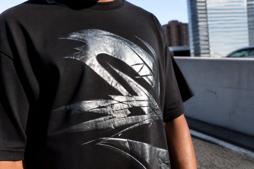 Close-up of a heavyweight cotton streetwear tee with reflective graphic details catching light, showing the tactile texture and metallic sheen of the print in natural outdoor lighting
