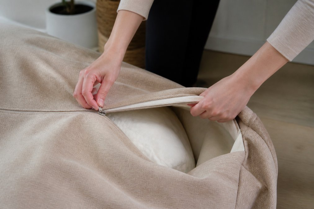 Gros plan des mains retirant délicatement une housse de pouf déhoussable par la fermeture éclair, montrant le processus d'ouverture et de séparation de la housse du rembourrage