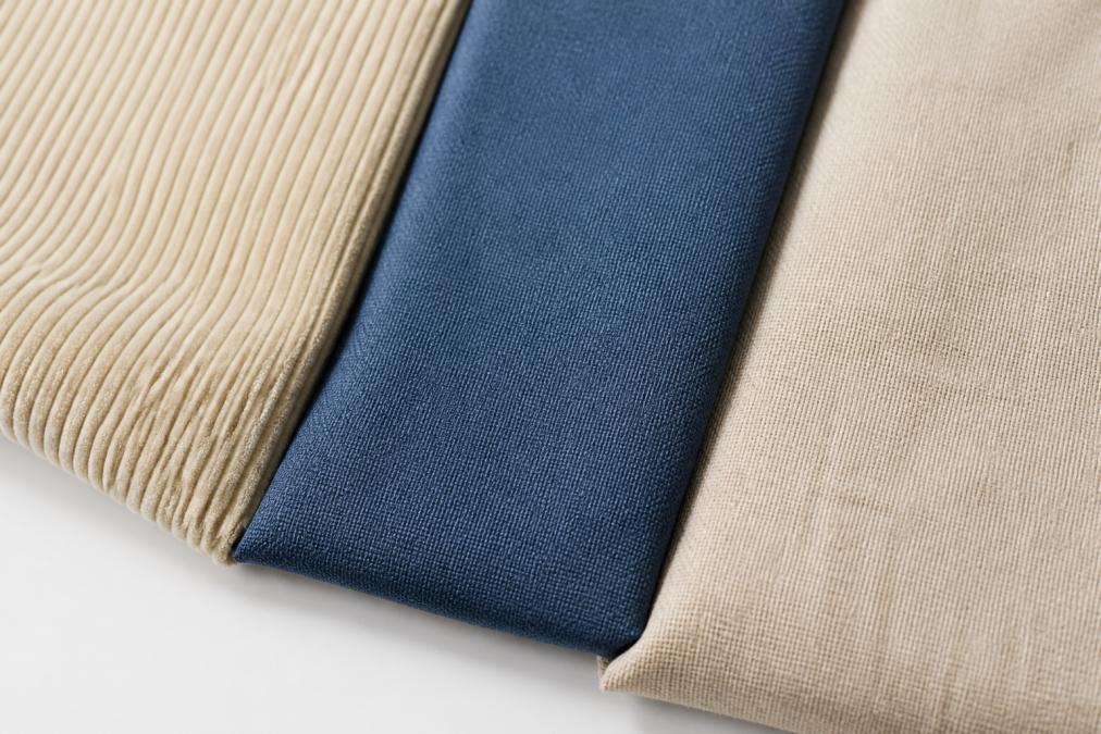 Détail photographié de plusieurs tissus de pouf (velours côtelé, polyester pur, mélange lin-coton) disposés côte à côte sur une surface blanche, montrant les textures, les finitions et les variations de couleur de chaque matériau
