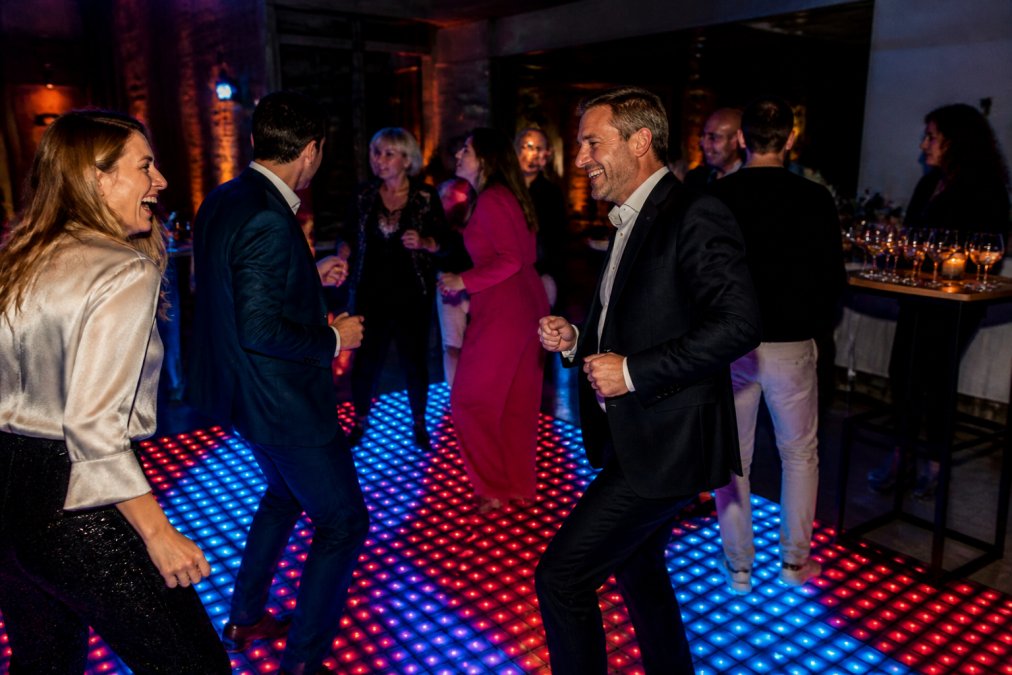 Groep zakenmensen van verschillende leeftijden dansend en lachend op een fel verlichte pixelvloer met rode en blauwe LED-effecten op een bedrijfsfeest, met champagneglazen op tafels op de achtergrond