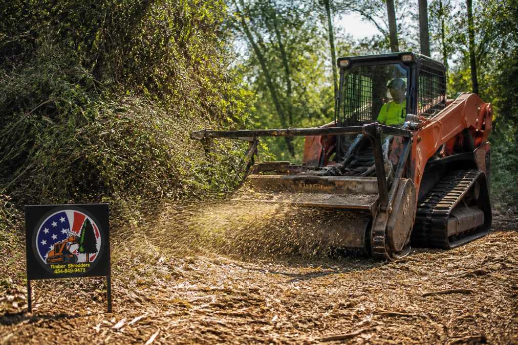 Professional forestry mulching equipment actively processing dense overgrowth into mulch on a rural property, with operator visible in equipment cab and processed material visible on-site