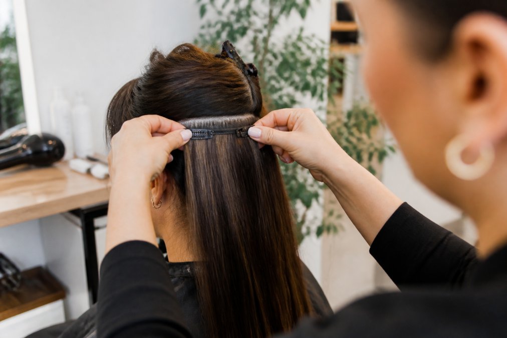 Professional demonstrating proper clip-in extension placement by carefully positioning silicone-lined clips into sectioned hair at the crown, showing hands securing extensions with precise grip in bright natural lighting
