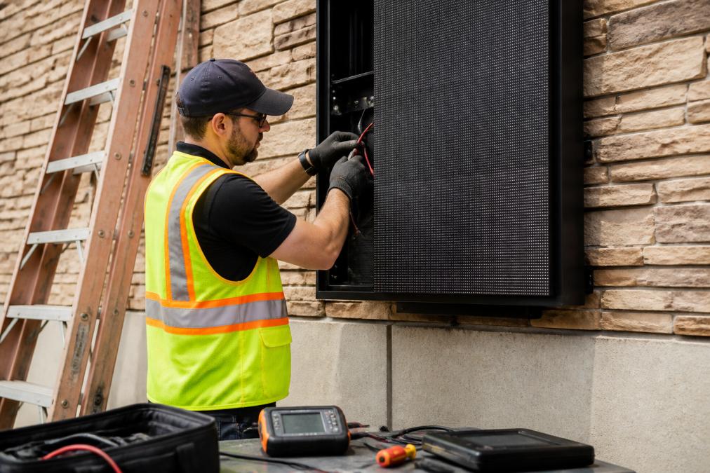 Technician in safety vest performing maintenance on large digital display sign mounted on exterior wall, checking cable connections and display functionality with diagnostic equipment in hand