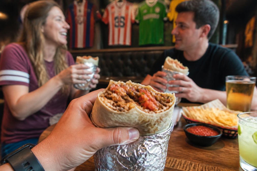 A close-up of a hand holding a large, foil-wrapped burrito with a bite taken out, revealing layers of spiced meat, rice, and vibrant red salsa, set against a rustic wooden table in a lively sports bar with blurred soccer jerseys in the background