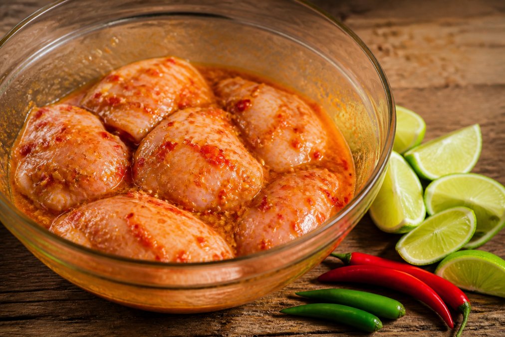 Raw chicken thighs in a glass bowl coated with a vibrant orange-red chili and citrus marinade, fresh lime wedges and chili peppers on a rustic wooden table nearby