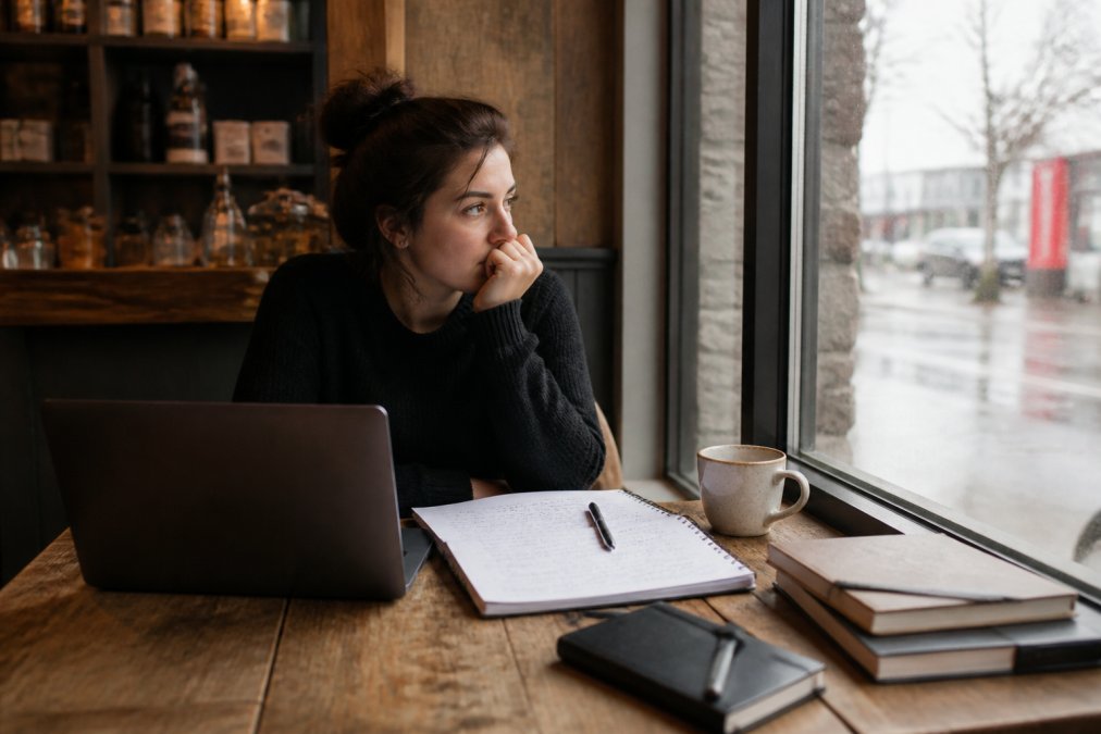 Person sitting alone at a coffee shop with a laptop nearby, looking thoughtful while working through notes, soft natural lighting from window