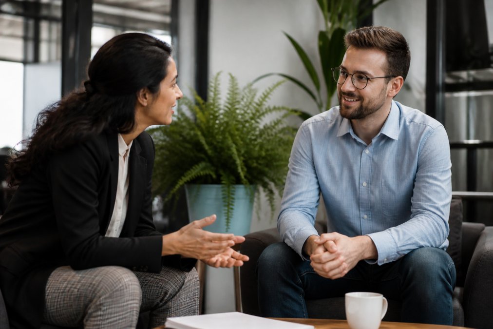 Two professionals having a relaxed conversation in a modern office setting, one leaning forward engaged, natural office lighting, plants in background
