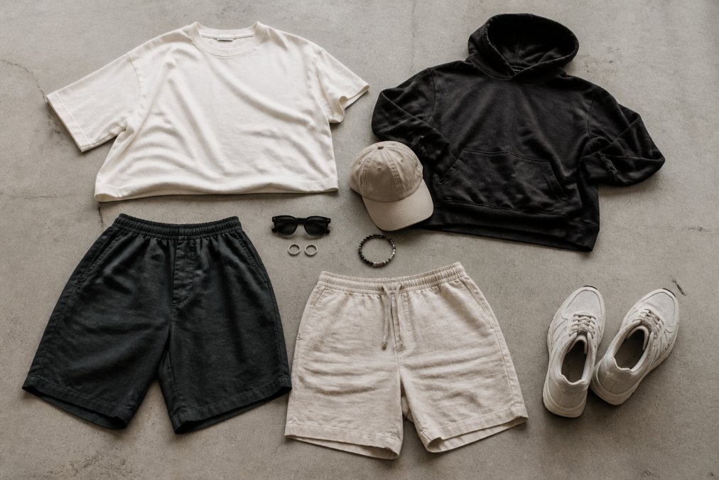 Flatlay van zomerse streetwear essentials: oversized t-shirt, bermuda shorts, linnen shorts, hoodie en accessoires netjes uitgelegd op een neutrale achtergrond