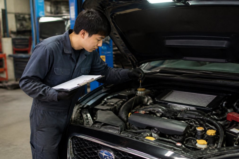 A mechanic in a clean shop inspecting a Japanese sedan's engine bay with a clipboard, checking fluid levels under hood light