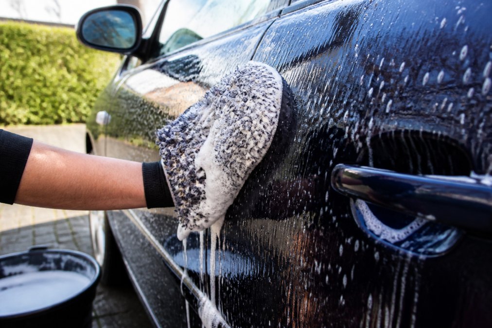 Close-up van een hand met een microvezel wash mitt die schuimende autoshampoo aanbrengt op een donkerblauwe autodeur, helder daglicht buiten