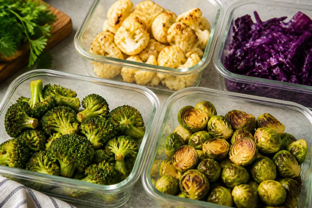 Overhead view of colorful meal prep containers with roasted broccoli florets, cauliflower, Brussels sprouts, and shredded purple cabbage arranged in glass storage bowls, natural kitchen lighting