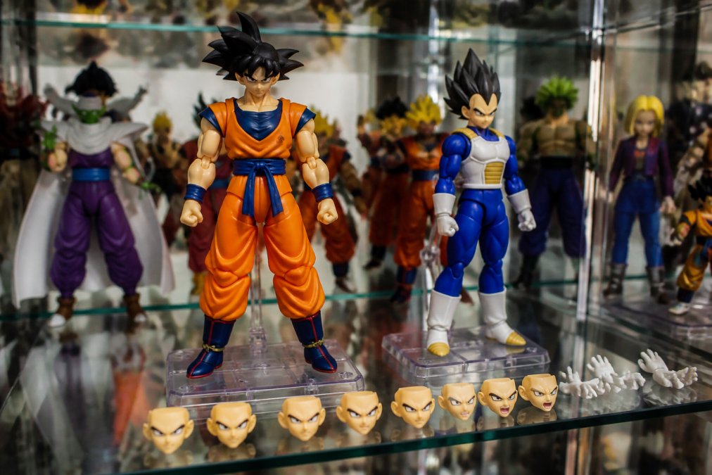 Close-up of a display shelf featuring articulated anime figures with detailed paint jobs and multiple interchangeable face plates arranged in a collector's case with museum-quality lighting
