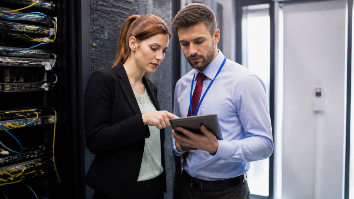 Business owner and IT consultant reviewing network infrastructure and security protocols on tablet in modern server room with natural lighting