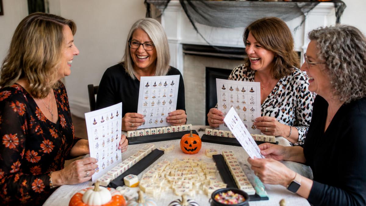 A group of women at a Mahjong table holding printed Halloween-themed game sheets, laughing together, with colorful tiles and small pumpkins on the table