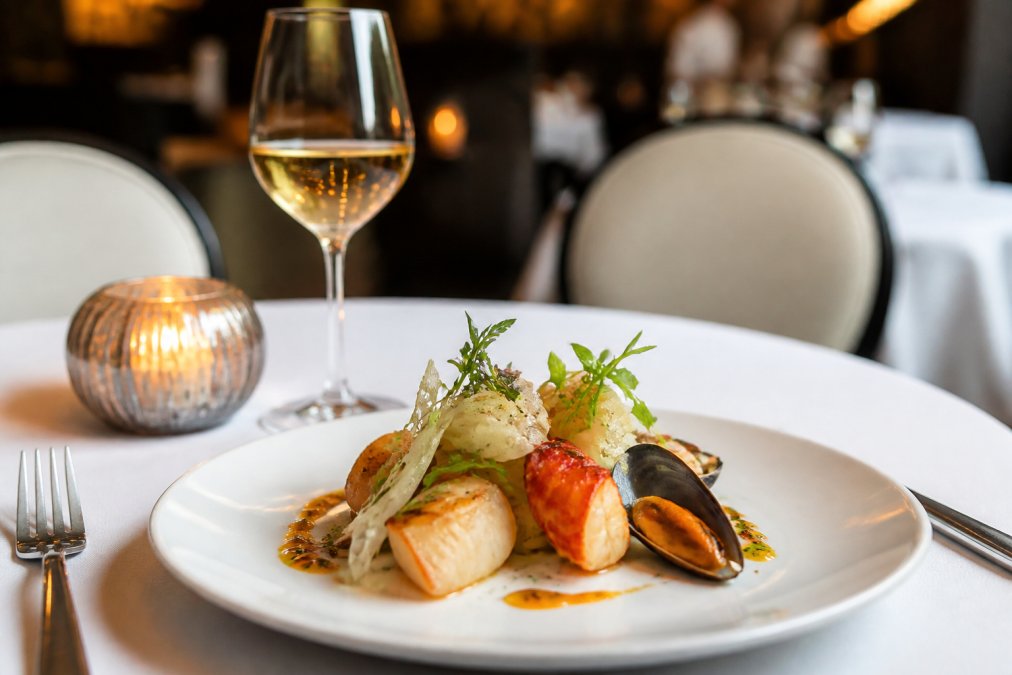 Elegantly plated seafood dish with fresh local ingredients, delicate garnishes, and wine glass on white tablecloth with soft candlelight in upscale restaurant setting