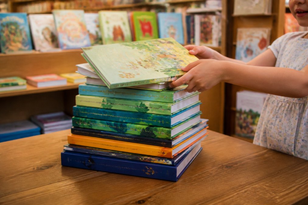 A colorful stack of new hardcover children's books displayed on a wooden table in a bright, cozy bookstore, with a child's hands reaching for one