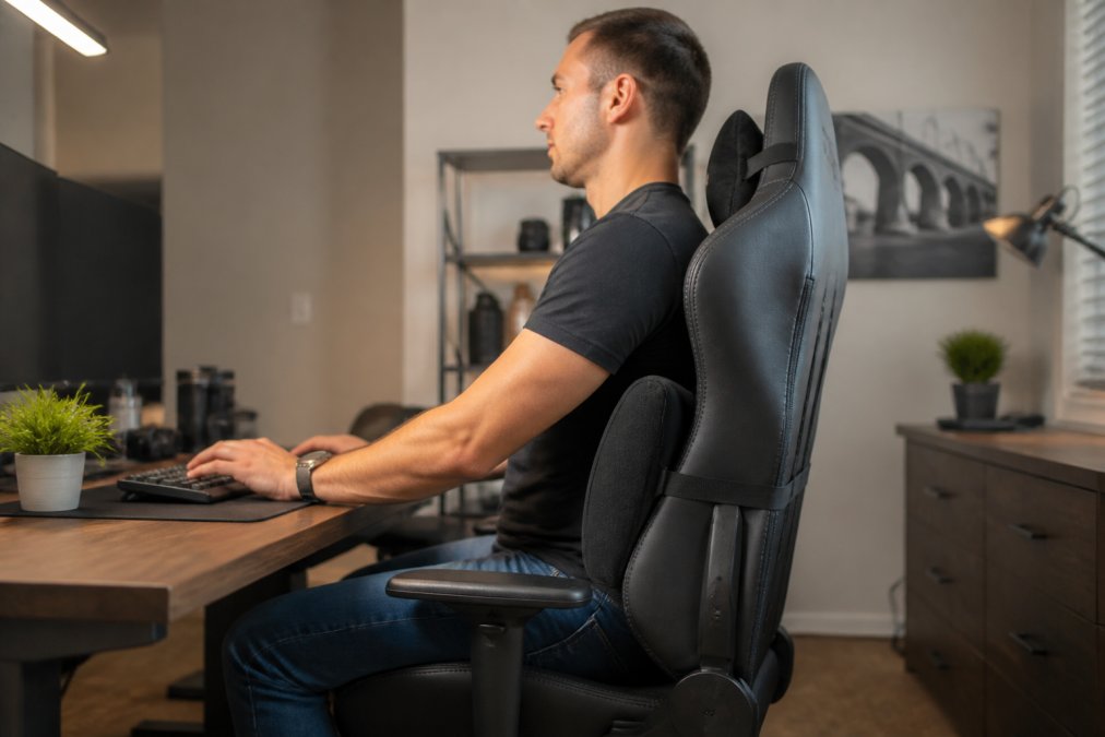 A person sitting upright in a tall-backed gaming chair at a dual-monitor desk, lumbar support cushion visibly pressing against the lower back, soft overhead lighting in a home office setting