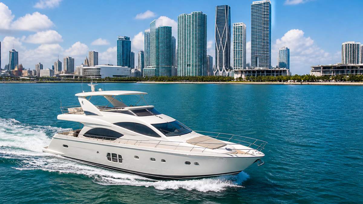 Aerial view of a white luxury motor yacht cruising Biscayne Bay in Miami with the downtown skyline behind it, clear blue water and bright midday sun