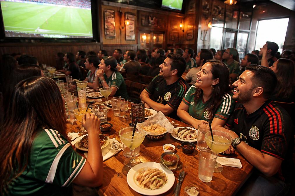 A lively group of friends in team jerseys gathered around a long table in a warm sports bar, plates of tacos and margarita glasses on the table, eyes fixed on a large screen showing a soccer match