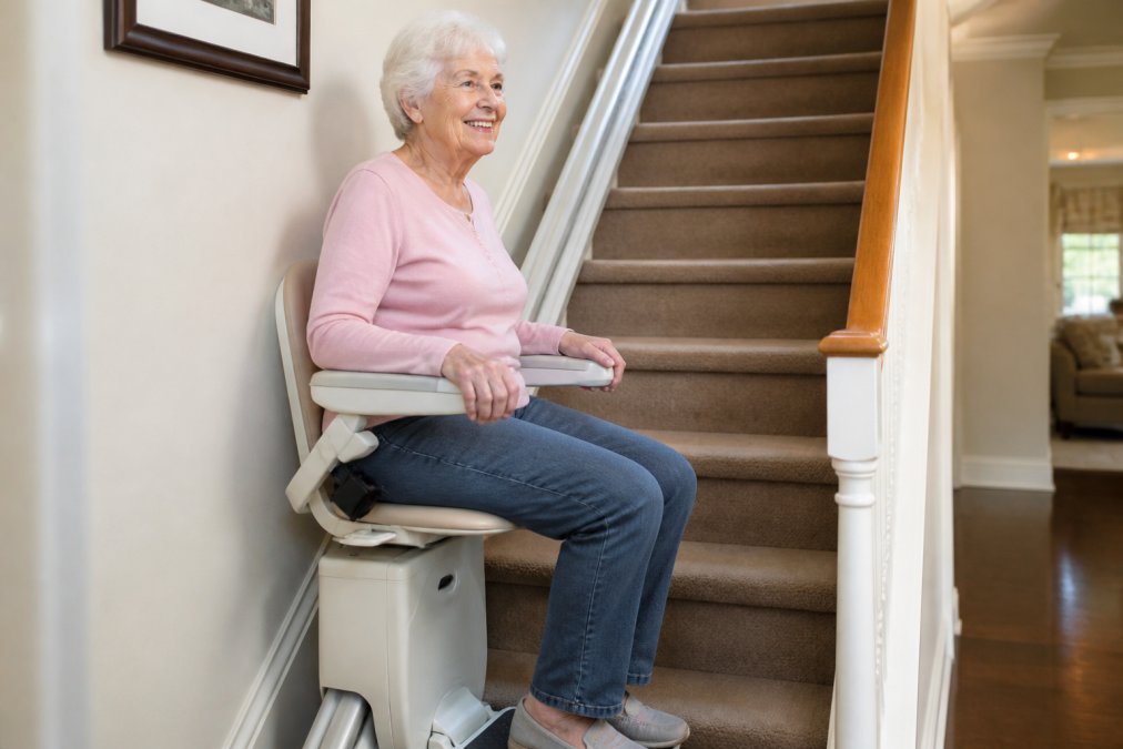 Older adult comfortably seated in a modern stairlift with visible armrests and safety features, ascending a residential staircase in a well-lit home interior