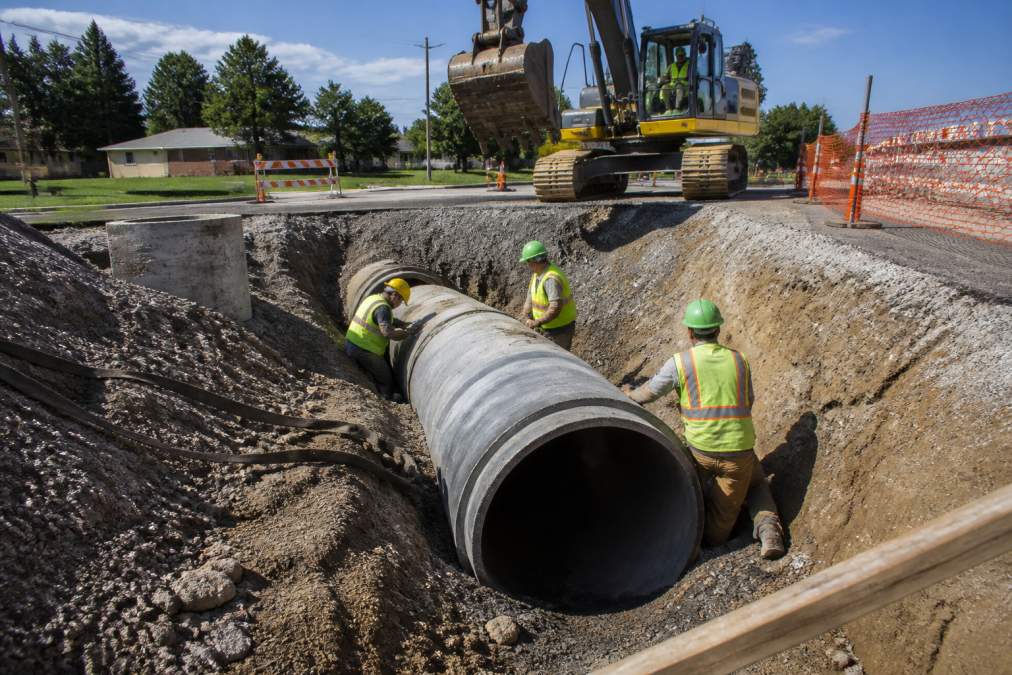 Professional construction crew installing large diameter water main pipe in deep excavation trench, with safety barriers, heavy equipment, and blue sky visible in background