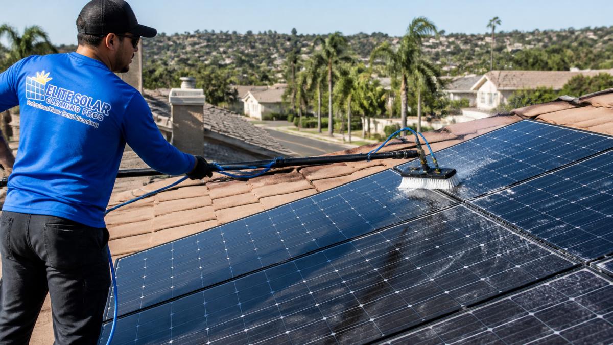 Professional technician using water-fed pole to clean solar panels on residential rooftop, clear panels visible in morning sunlight with San Diego neighborhood in background