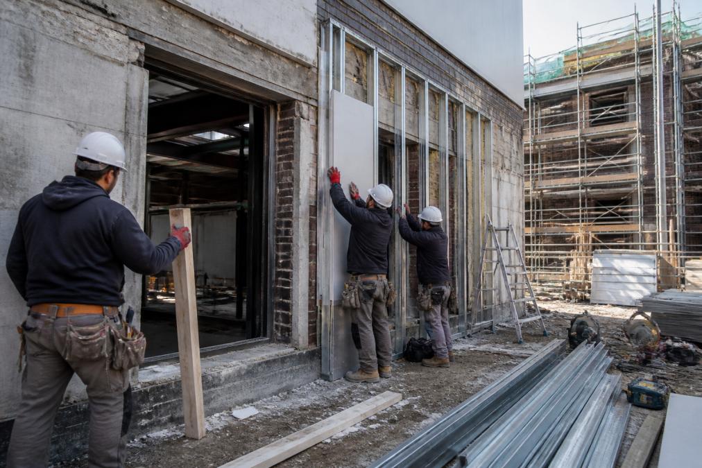 Bouwteam op een bedrijfspand met bouwhelmen en gereedschap, werkend aan gevelisolatie en structurele aanpassingen, helder daglicht op de werkplek