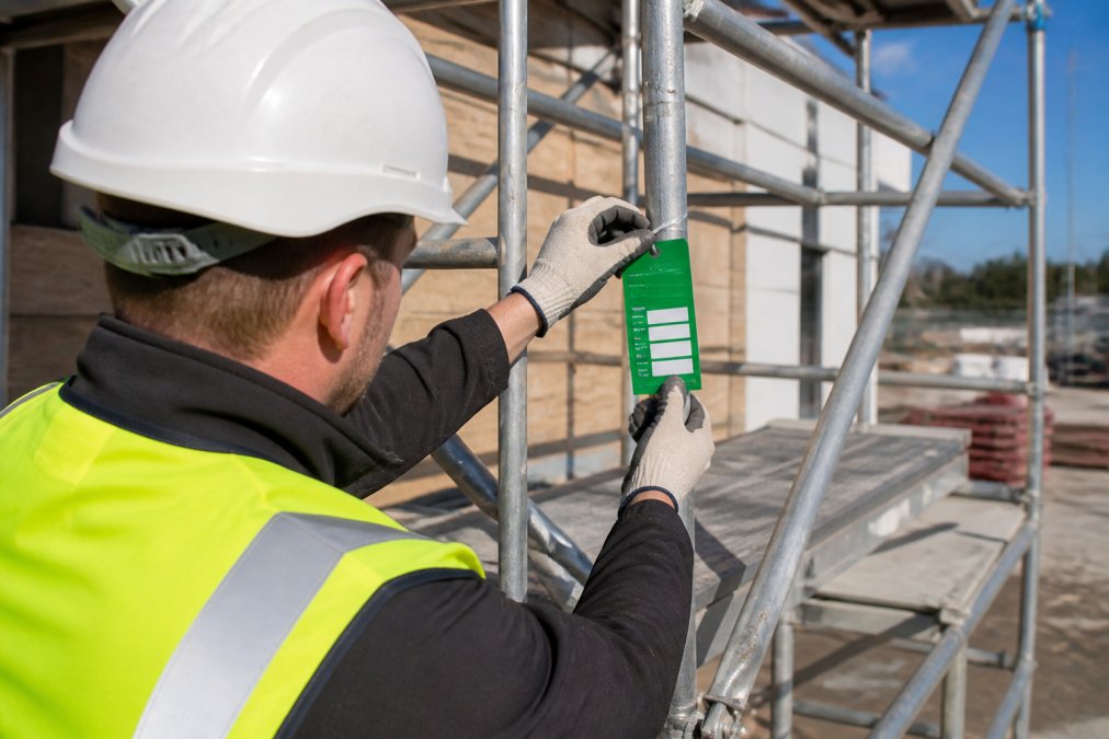 Close-up of a worker's gloved hand attaching a completed green scaffold safety tag to the frame of an aluminium tower scaffold, bright daylight on a construction site
