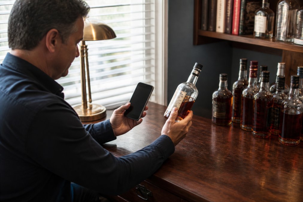 Bourbon collector sitting at a desk with multiple bottles arranged nearby, scanning a bottle label with a smartphone to log it into a tracking app