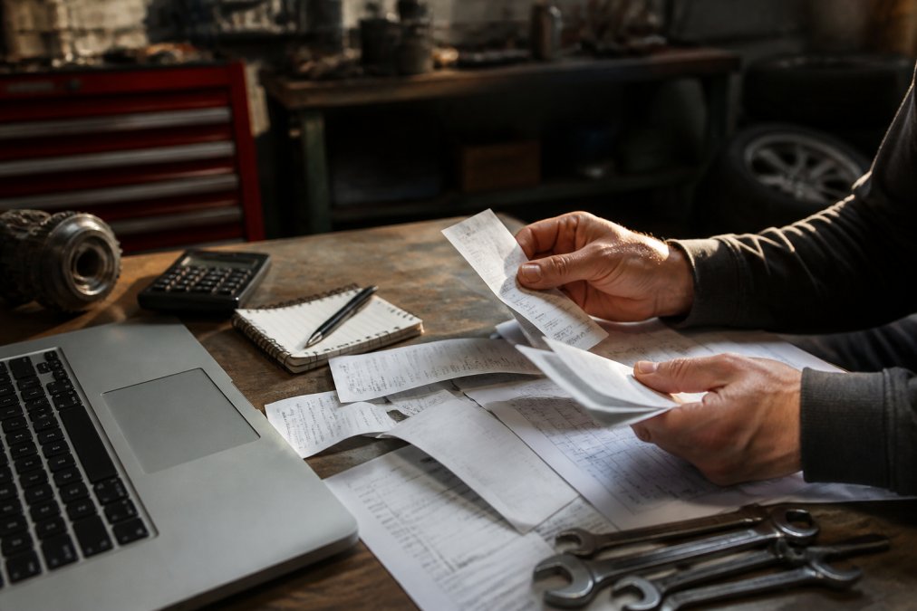 Close-up of hands organizing receipts and invoices on a work desk with a laptop displaying a spreadsheet, tools and car parts visible in the background under warm lighting