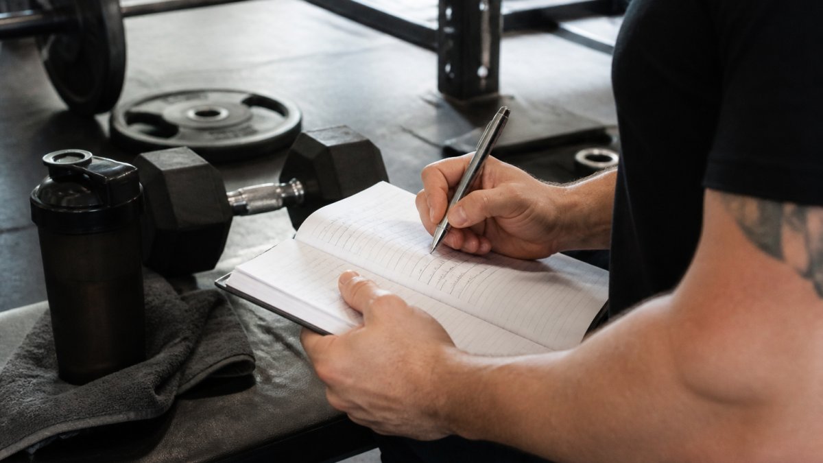 Fitness enthusiast writing detailed notes in a physical workout log at a gym station with dumbbells and barbells visible in the background, natural gym lighting