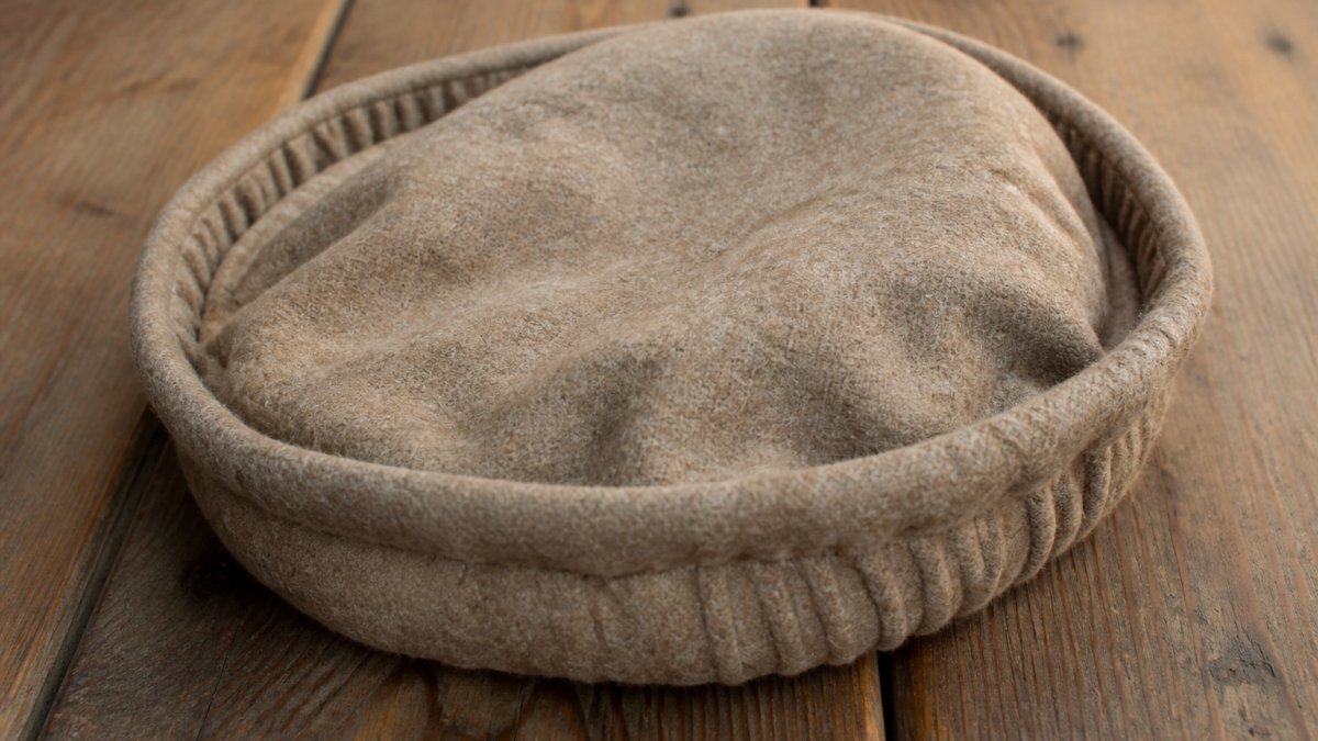 A close-up photograph of a hand-stitched wool pakol hat in light brown, resting on a wooden surface, with visible rolled brim and soft round crown, natural daylight