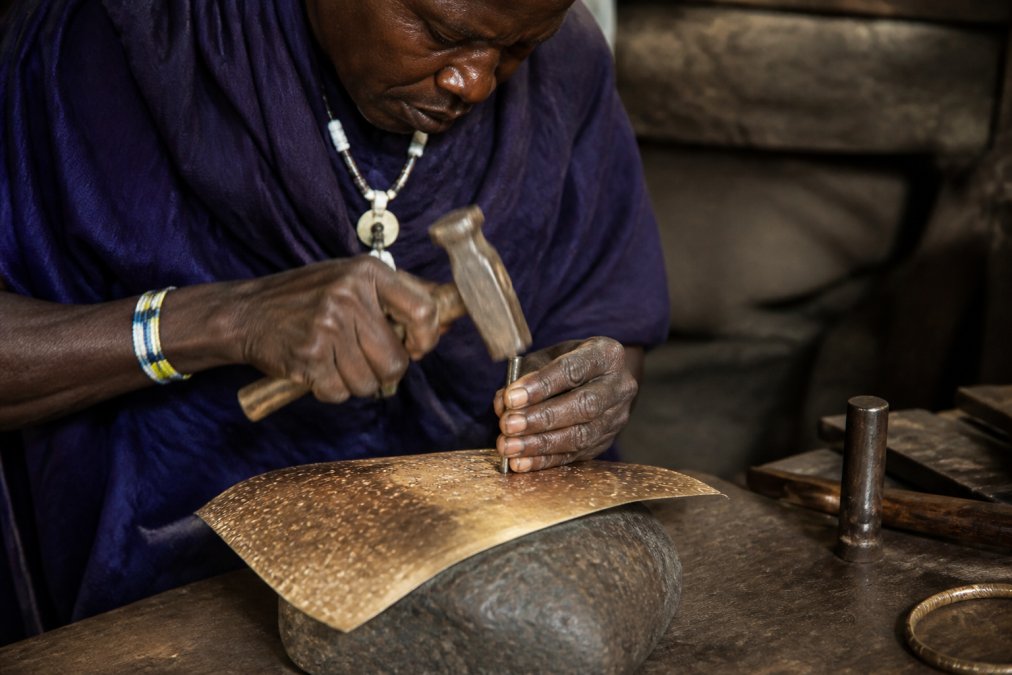Close-up of an artisan's hands using specialized hammers and tools to hand-hammer and shape gleaming metal sheets, with focused concentration and traditional metalworking techniques visible in the detailed work