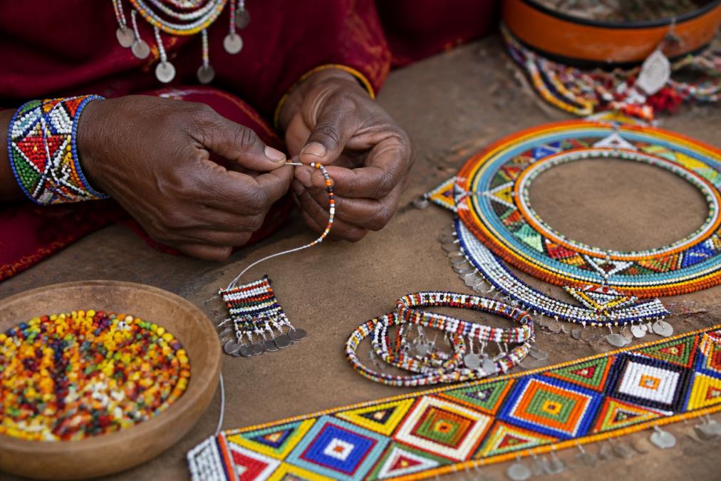 Artisan's hands carefully stringing colorful traditional beads and materials by hand, with finished beaded jewelry pieces displaying intricate geometric patterns arranged nearby on a work surface