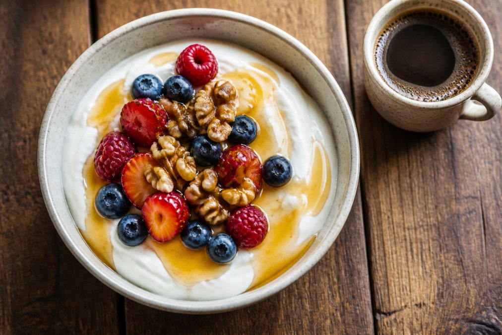 Close-up overhead shot of a bowl of thick Greek yogurt drizzled with golden honey and topped with walnuts and fresh berries, sitting on a rustic wooden table with a small cup of Greek coffee nearby
