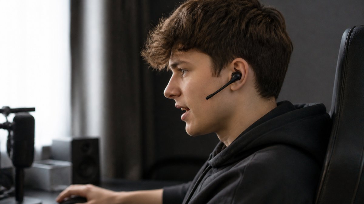 Gamer med trådløse ørepropper sitter ved PC-en med fokus på skjermen, mikrofonen tydelig synlig på øreproppen mens han snakker