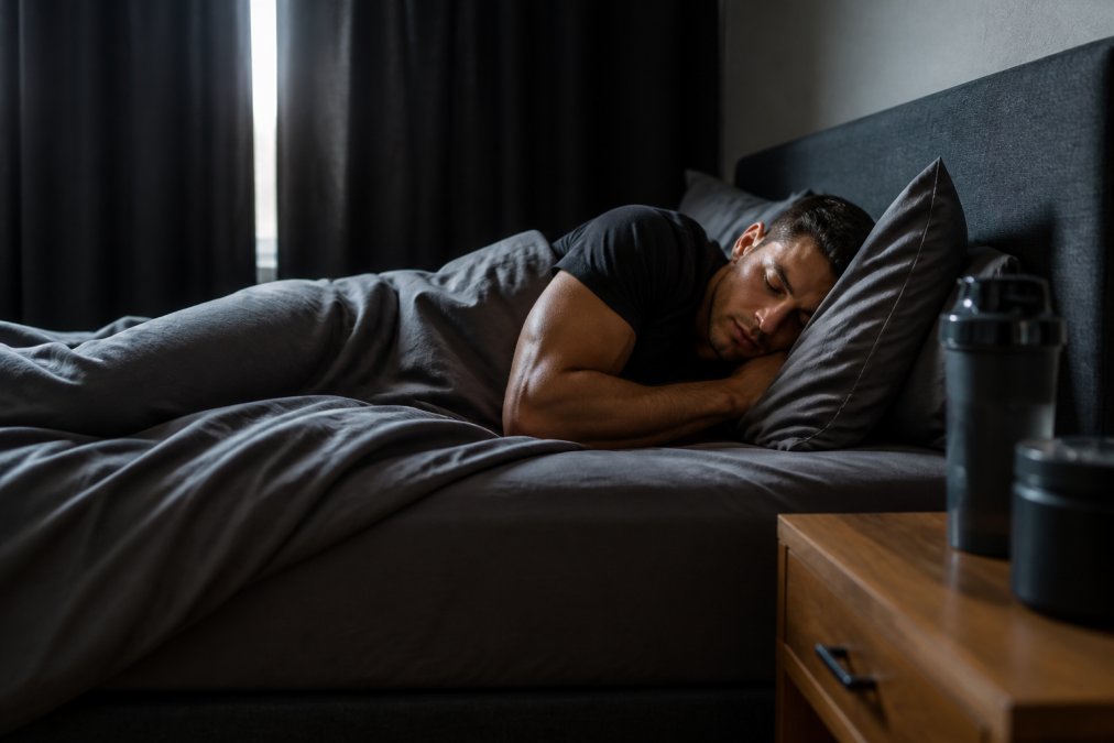 Person sleeping peacefully in a darkened bedroom with soft natural light filtering through blackout curtains, appearing deeply relaxed and well-rested