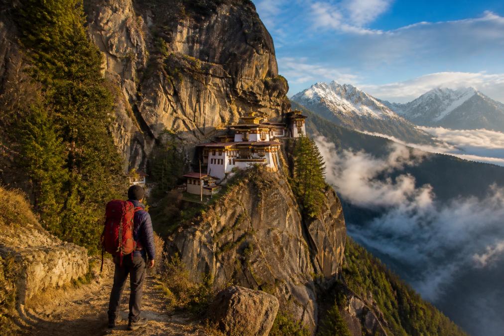 A traveller standing on a mountain pathway overlooking Tiger's Nest Monastery perched dramatically on a cliff face, with mist-covered valleys and snow-capped Himalayan peaks in the background, golden morning light illuminating the white fortress structure