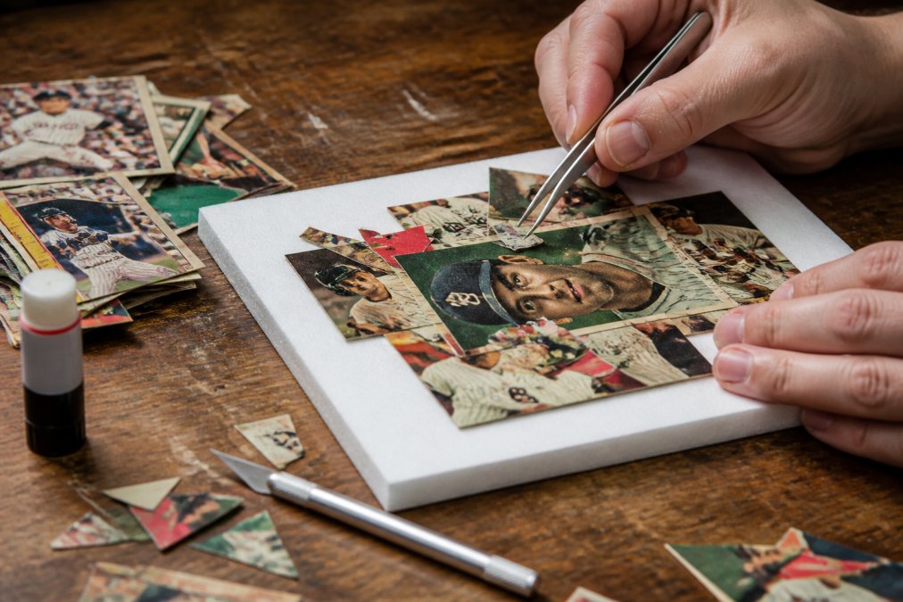 Close-up of hands using precision tweezers to layer cut baseball card fragments onto a foam board, with an X-Acto knife, glue stick, and a pile of vintage cards on a wooden workbench