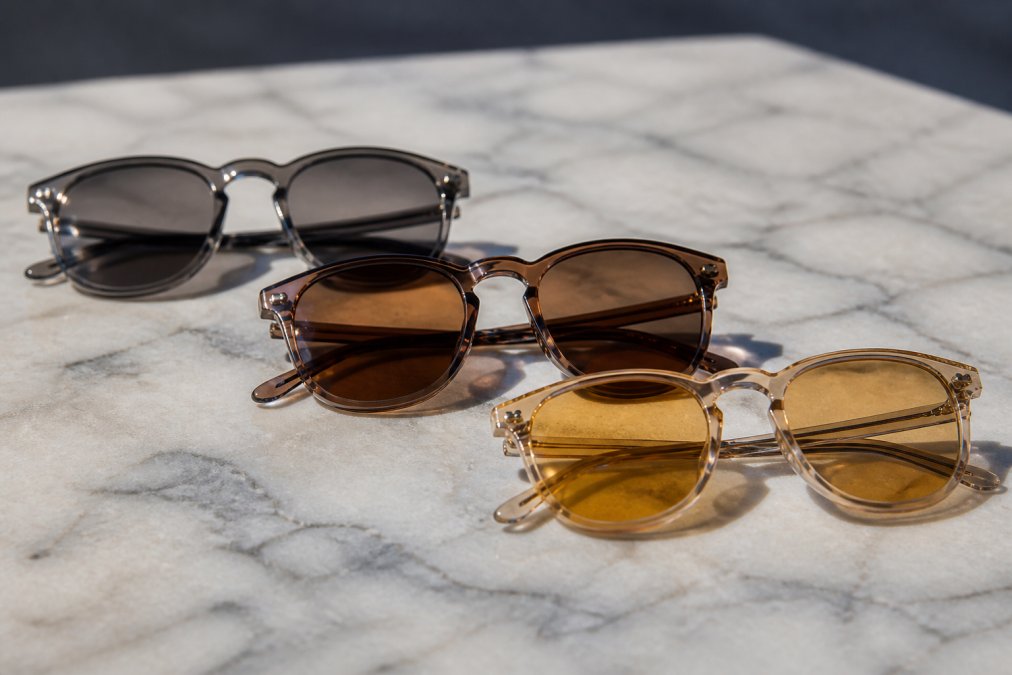 Close-up of multiple pairs of eyeglasses with different lens tints including gray, brown, and amber displayed side-by-side in natural daylight on a neutral background
